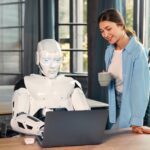 A photo of a robot and a woman in front of a computer.